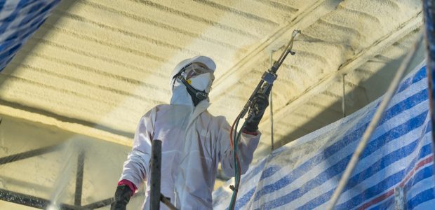 A professional in full gear and in action of applying a spray foam roofing system which can help improve a roof’s structural integrity by locking its components in place