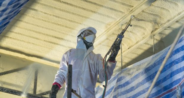 A professional in full gear and in action of applying a spray foam roofing system which can help improve a roof’s structural integrity by locking its components in place